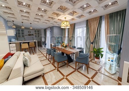 Living room of a large country house with a large table for eight seats. Interior with beautiful curtains. Chandelier lights on. Square ceiling.