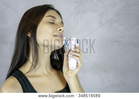 Young Pretty Woman Doing Skin Care Procedure. Cleaning Face With Machine On Grey Background