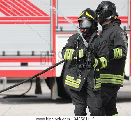 Two Firemen With Hose Hydrant In Action And Fire Engine On Background