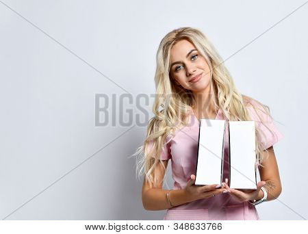 Blonde Girl In Pink Medical Uniform And Jewelry Posing Isolated On White. She Smiling, Holding Packa