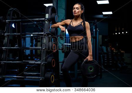 Front View Of Fitnesswoman With Braids Posing Near Stand With Sports Equipment And Holding Weights. 
