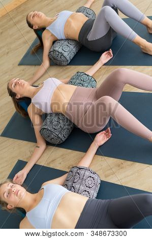 Restorative Yoga With A Bolster. Group Of Three Young Sporty Attractive Women In Yoga Studio, Lying 