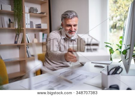 Architect With Model Of House Sitting At The Desk Indoors In Office, Working.