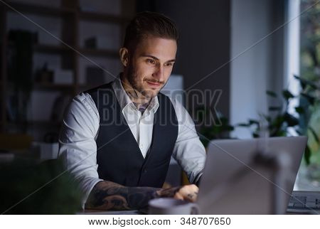 A Businessman With Laptop Sitting At The Desk Indoors In Office, Working Late.