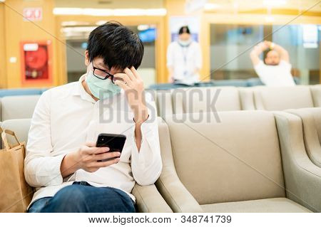 Male Asian Patient Wearing Surgical Mask Using Smartphone At Waiting Area In The Hospital Or Medical