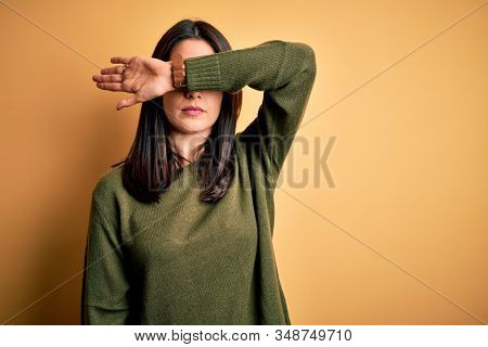 Young brunette woman with blue eyes wearing green casual sweater over yellow background covering eyes with arm, looking serious and sad. Sightless, hiding and rejection concept