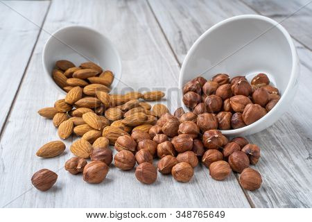 Scattered Hazlenuts And Almond Seeds With White Bowls On Wood Table