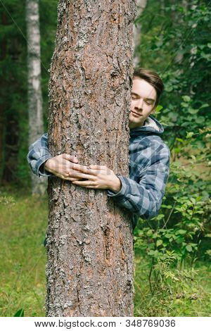 Teenager boy hugging pine tree in a summer forest leaning to tree trunk with closed eyes embracing the nature