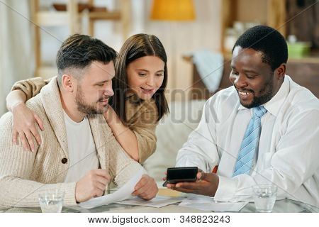 Adult smiling black man with calculating machine showing young couple their interest rate