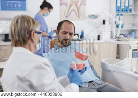 Senior Woman Doctor Showing Artificial Jaw Model To Patient With Toothache Discussing Carier Treatme