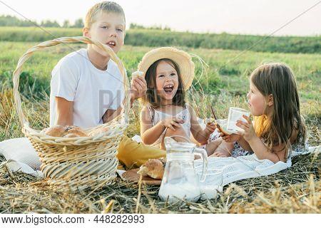 Portrait Of Joyful Boy And Two Girls Sitting On Blanket In Field, Having Picnic, Eating Bread, Drink