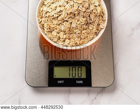 Raw dry oatmeal in a bowl on a digital kitchen scale. The scales displaying 100g of grits without tare. Modern electronic device for measurement the food weight for dieting and cooking. Top view.