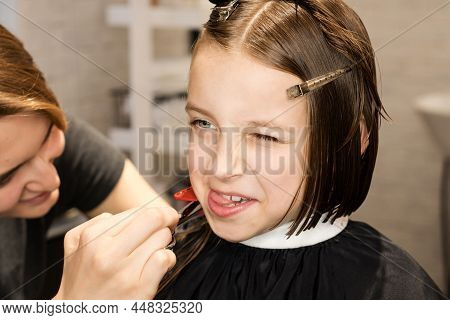Gorgeous Happy Short-haired Little Girl Makes Grimaces In Barbershop In Process Of Cutting Hair In B