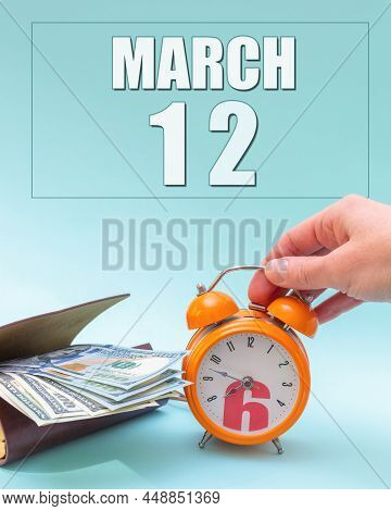 March 12th. Hand Holding An Orange Alarm Clock, A Wallet With Cash And A Calendar Date. Day 12 Of Mo