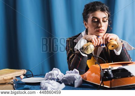 Upset Newswoman Sitting With Handset Near Vintage Typewriter And Crumpled Paper On Blue Background