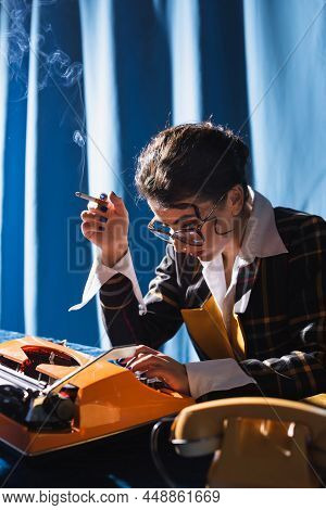 Stylish Newswoman In Eyeglasses Holding Cigarette While Working At Typewriter On Blue Background