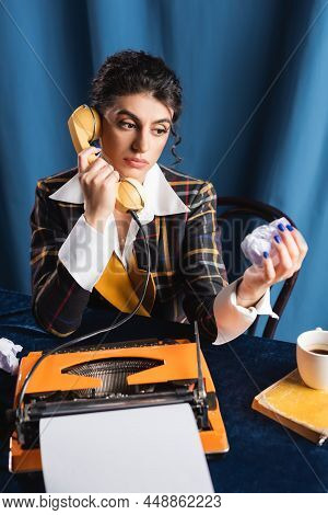 Pensive Newswoman With Crumpled Paper Talking On Telephone Near Typewriter On Blue Background