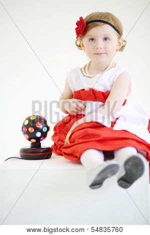Little pretty girl sits near ball-shaped lamp on white background.