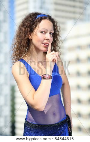 Woman with curly hair in a blue suit and a scarf on hips holding a finger near lips against the background of a multistory residential complex