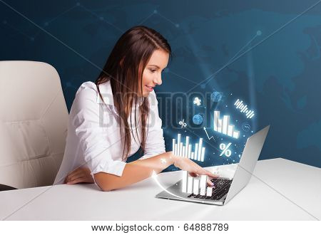 Pretty woman sitting at desk and typing on laptop with diagrams and graphs comming out