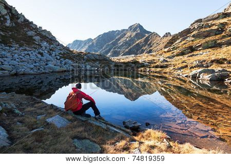 Male hiker takes a rest sitting next a mountain lake. Autumn sunny day. West italian Alps, Oropa, Piemonte, Italy, Europe.
