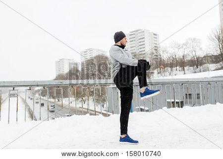 fitness, sport, people, exercising and healthy lifestyle concept - young man stretching leg and warmig up on snow covered winter bridge