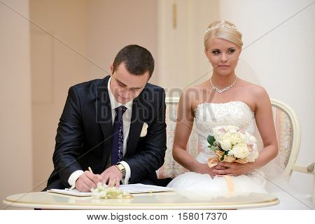 Beauty bride and handsome groom are registering the marriage. Wedding couple is marrying. Beautiful model girl in white dress and in lace veil. Man in suit. Female and male portrait. Cute lady and guy