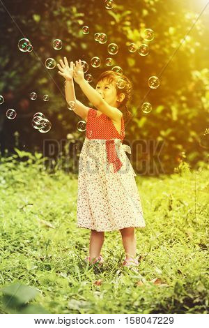 Asian lovely girl playing on green grass at park . Happy child reaching for soap bubbles on blurred nature background. Outdoor at the daytime with bright sunlight. Warm tone and vintage effect style.