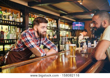 Happy attractive young barman giving glass of beer and talking to african american young man 