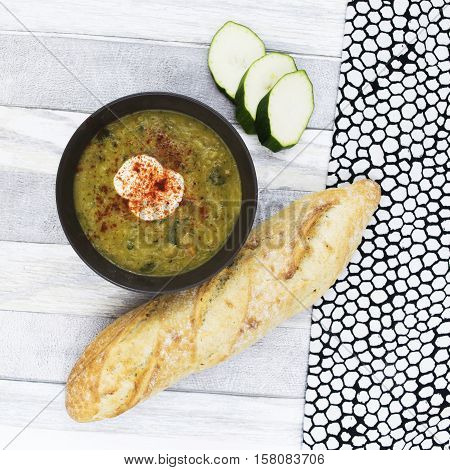 Courgette soup with sour cream in black bowl . Sliced zucchini and long fresh baguette on the white wooden background. Top view