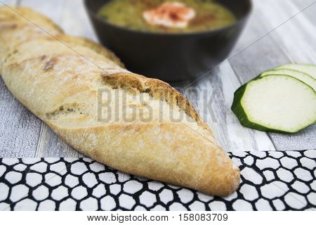 Courgette soup with sour cream in black bowl . Sliced zucchini and long fresh baguette on the white wooden background