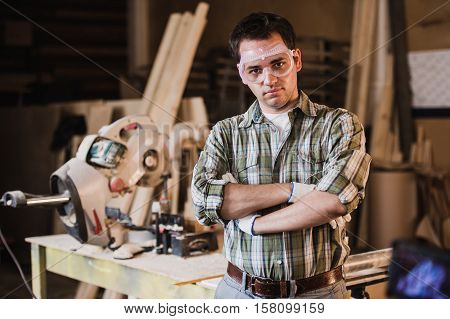 Handsome carpenter in protective glasses is looking at camera and smiling while standing near his wooden object in the workshop.