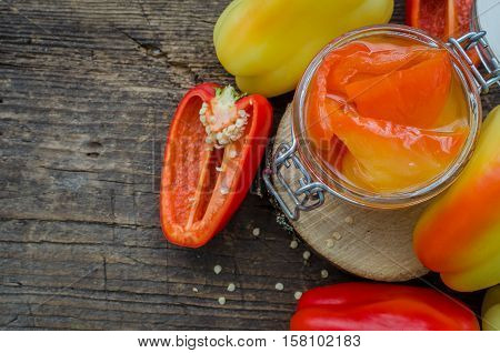Bell pepper preserved in a glass jar with fresh peppers on old wooden background. Homemade marinated in oil red pepper. Glass jar with conserved roasted yellow and red paprika. Top view. Copy space.