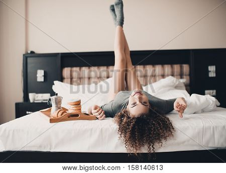 Beautiful smiling girl 20-24 year old having fun in bed with breakfast on wooden tray. Wearing knitted socks and t-shirt. Looking at camera.