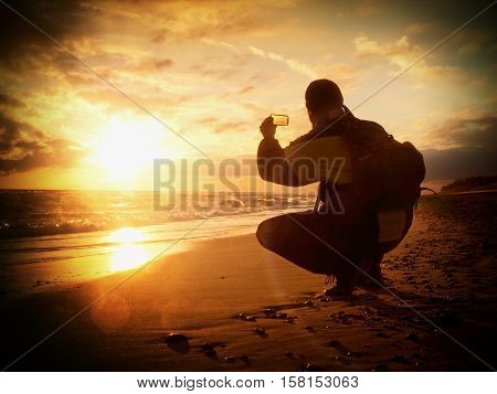 Man Takes Photo Of  Evening Sea. Hiker With Backpack With Phone In Hand. Tourist Squatting