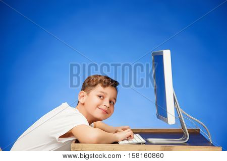 School-age boy sitting in front of the monitor laptop at blue studio backgroubd