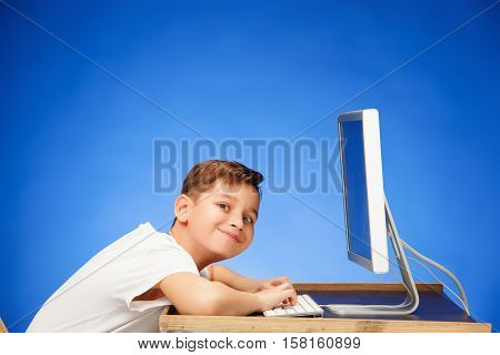 School-age boy sitting in front of the monitor laptop at blue studio backgroubd