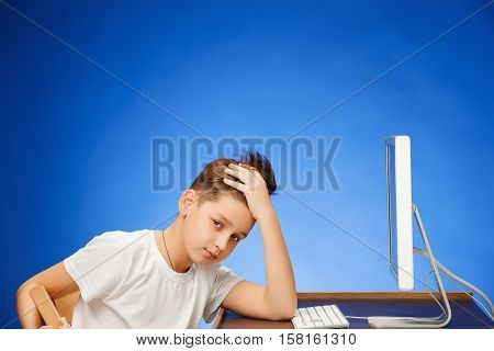 School-age tired boy sitting in front of the monitor laptop at blue studio backgroubd