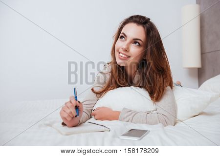 Smart pensive girl lying in bed and making notes in notebook at home