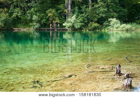 Small figures of tourists walking along a hiking path across the lake with emerald transparent water. Plitvice lakes national park Croatia