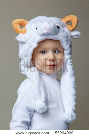 Cute adorable pretty Caucasian baby toddler with large blue eyes wearing a sheep hat hood with yellow horns on top and white shirt standing on a light background looking into the camera New Year concept studio