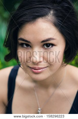 Closeup portrait of a beautiful young asian japanese girl woman with freckles with black short haircut looking into the camera in garden with green leaves