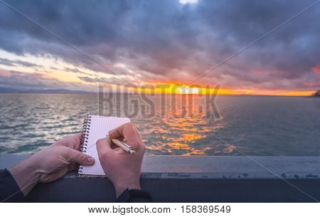 Writing on a notebook at sunset - Conceptual image with a man writing in a spiral notebook while the sun sets down over the Bodensee lake.