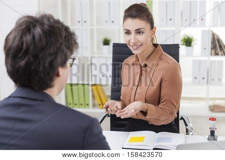 Smiling office employee in brown blouse is talking to her colleague wearing glasses and sitting with his back to the camera. Overt the shoulder shot.
