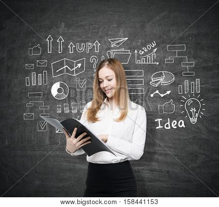 Smiling businesswoman with a black folder is standing near a blackboard with startup idea sketch. Concept of business foundation