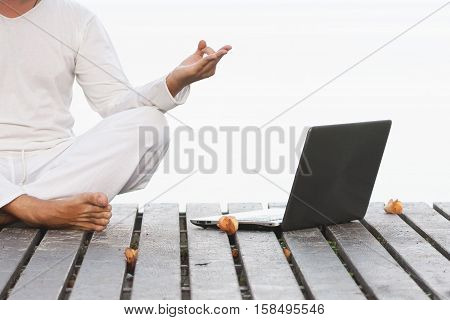 Caucasian man in white clothes meditating yoga with laptop on wooden pier