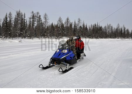 Two men ride on a snowmobile on the frost river in the winter forest