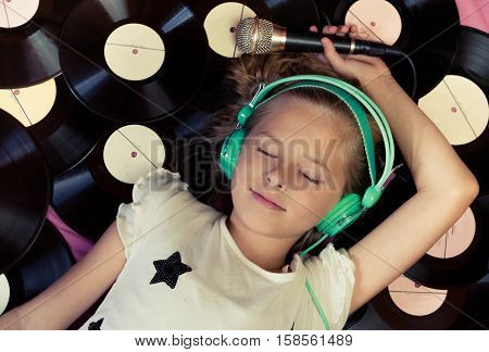 Beautiful girl lying next to vinyl records holding a microphone and listening music on headphones.Top view.