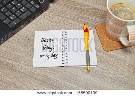 Open notebook with text"Do new things every day" and a cup of coffee on wooden background. Top down view