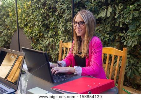 Young enterprising girl working with laptop on the terrace of her house. She is wearing glasses and she is blonde.